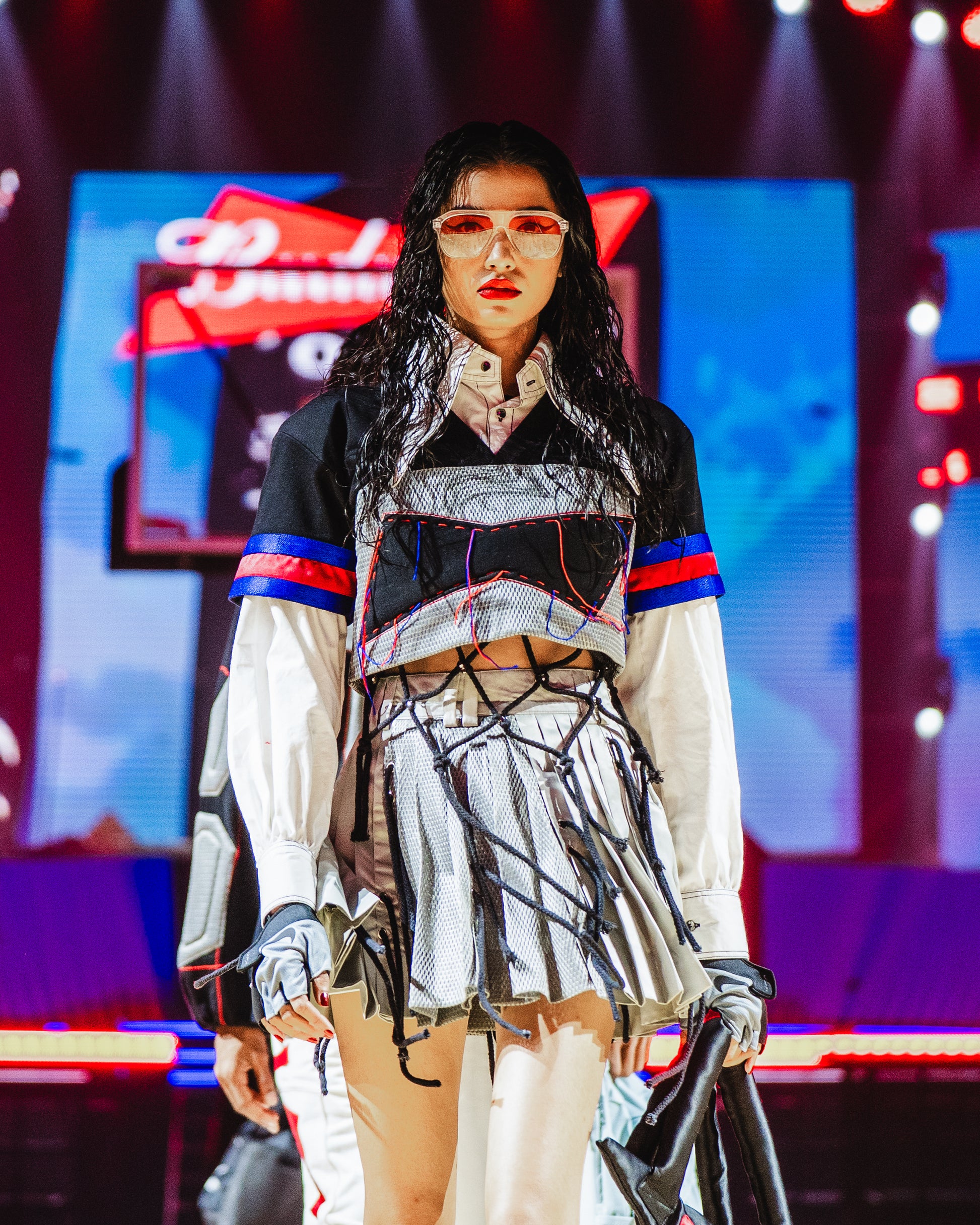 Model walking on a runway at a BUDXNBAHOUSE fashion show wearing black knotted layering top.