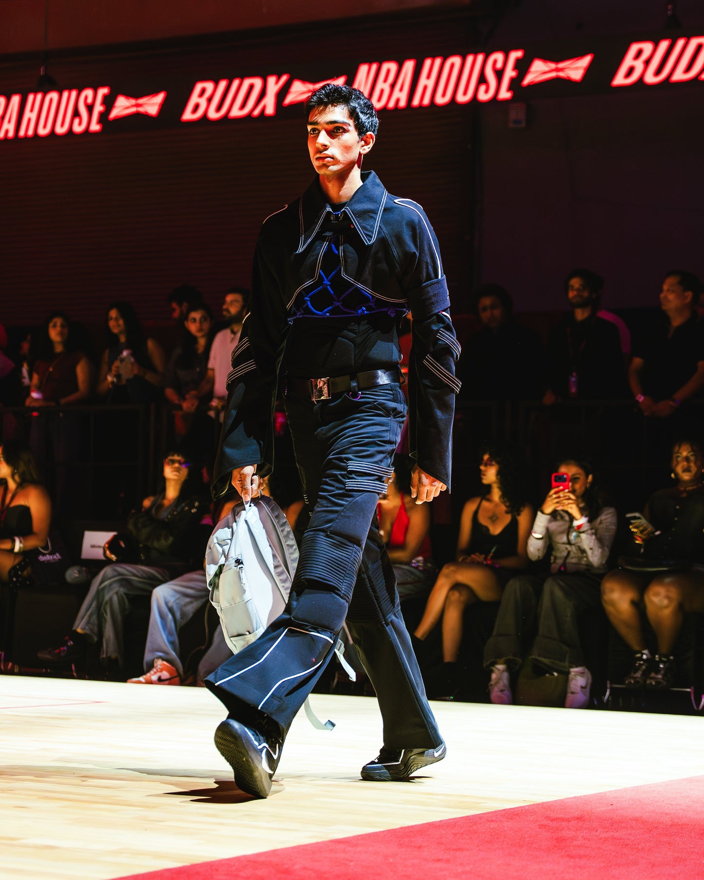 Model walking on a runway at a BUDXNBAHOUSE fashion show wearing blue knotted layering top.